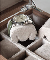 A wristwatch with a silver case and green strap is placed on a cushioned pillow inside a TAWBURY Grove Watch Box Pillow - X-Small in Beige.